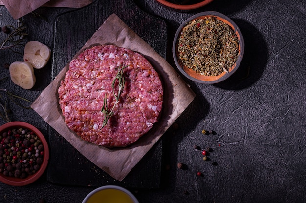 Rohes Schweinefleisch-Hamburgerkotelett mit Gewürzen auf dunklem Hintergrund. Heimproduktion von Halbzeugen. Einfaches Essen.