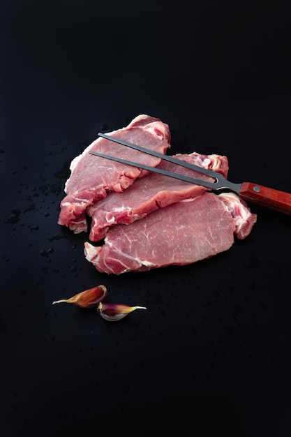 Rohes Schweinefleisch zum Grillen und Fleischgabel auf dunklem Hintergrund Stücke rohes Roastbeeffleisch zum Grillen Frisches Rindfleisch zum Grillen