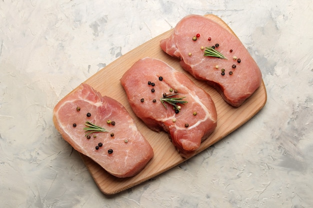 Rohes Schweinesteak auf dem Brett, Fleisch und Zutaten zum Kochen, Gewürzkräuter und Gemüse auf hellem Hintergrund. Ansicht von oben