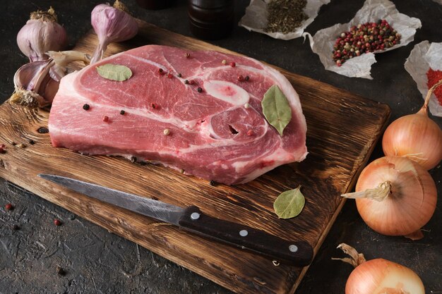 Rohes Schweinesteak auf einem alten Schneidebrett, Messer, Gewürze und Zutaten liegen nebeneinander.