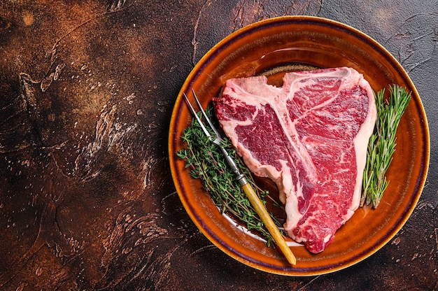 Rohes T-Bone-Rindfleisch Steak mit Kräutern auf einem Teller. Dunkler Hintergrund. Ansicht von oben. Platz kopieren.