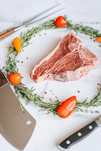 Rohes Tbone-Steak in der Mitte eines Weihnachtskranzes aus Rosmarin-Thymian-Kirschtomaten und Paprika auf einem weißen Marmoroberflächen-Weihnachtsessen-Konzept