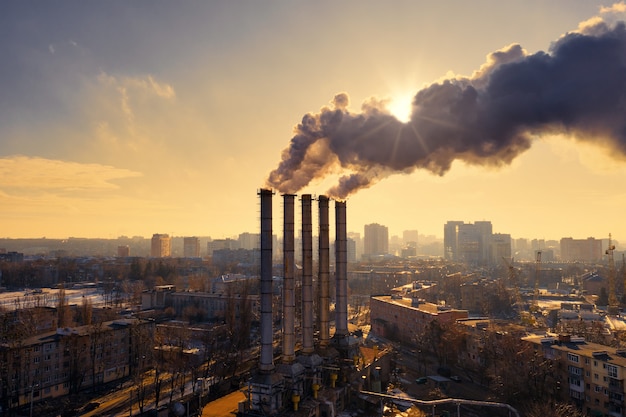 Rohre einer Industriefabrik mit schwarzem Rauch, der die Sonne während des gelben Sonnenuntergangs im Winter in der Stadt bedeckt