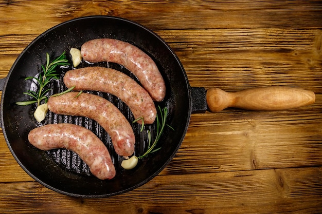 Rohwürste bereit zur Zubereitung mit Rosmarin-Knoblauch und Gewürzen in gusseiserner Grillpfanne auf Holztisch Draufsicht