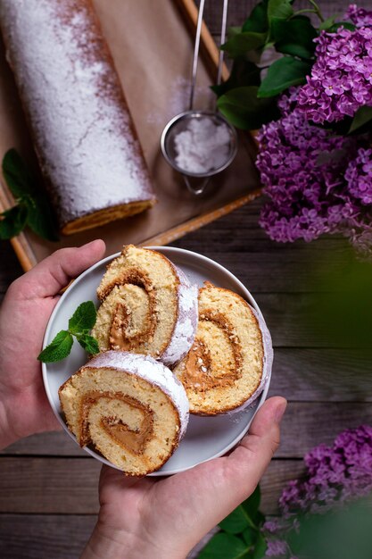 Rolinho de esponja caseiro com creme em um prato nas mãos sobre o fundo de uma mesa de madeira com flores da primavera