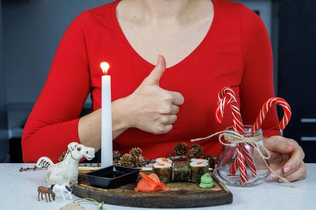 Rolinhos japoneses com peixe vermelho em uma tábua de madeira Uma garota com um suéter vermelho come sushi com pauzinhos Decoração de mesa com velas e doces Conceito de Natal