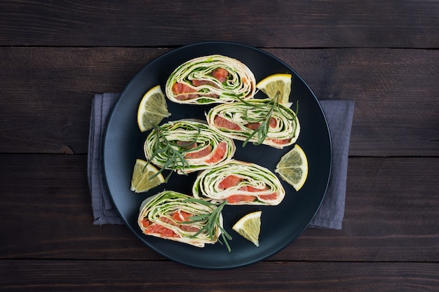 Rollen von dünnem fladenbrot und rotem gesalzenem lachs mit salatblättern auf einem schwarzen keramikteller, dunkler holztisch.