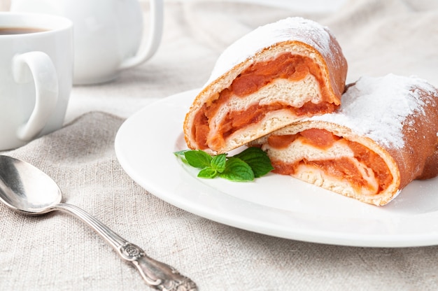 Rollo dulce con masa de levadura manzana espolvoreada con azúcar glas Cortada por la mitad Se encuentra en un plato blanco Decorado con una ramita de menta Hecho en casa En el fondo hay una tetera de porcelana blanca