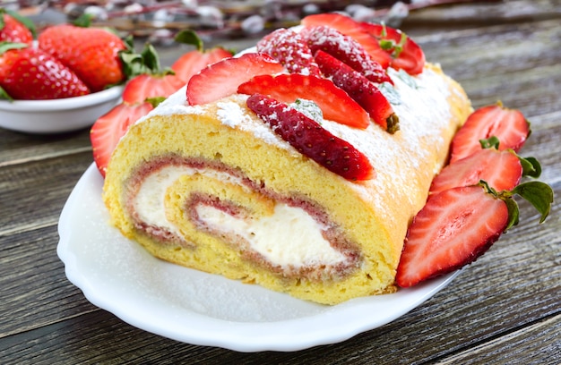 Rollo de galleta dulce con fresas y crema sobre una superficie de madera.