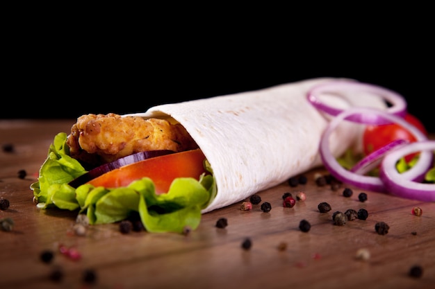 Rollo de pollo con lechuga tomate cebolla y pimiento sobre una mesa de madera y fondo negro.