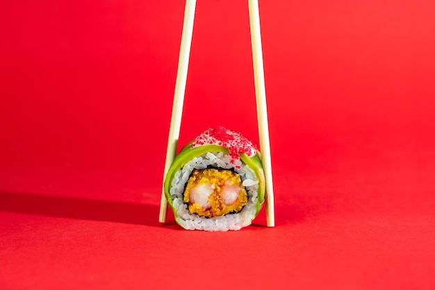 Rollo de sushi individual con aguacate de camarón y caviar de pez volador sobre fondo rojo con palos de madera Imagen creativa sobre fondo de color con sombra larga