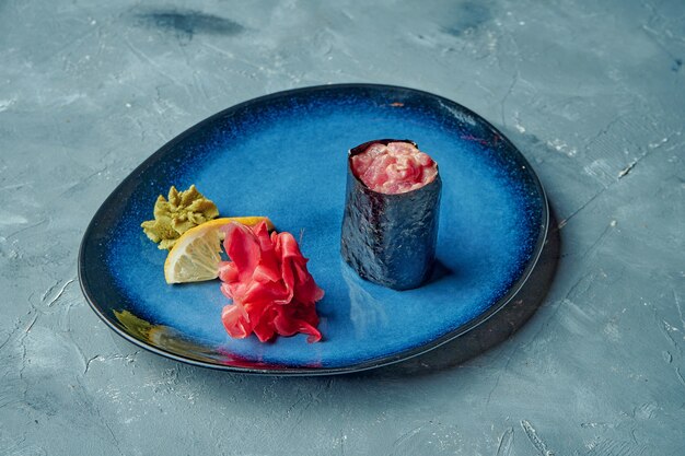 Foto rollo de sushi japonés con salsa picante y atún en una placa azul sobre una superficie gris. gunkan