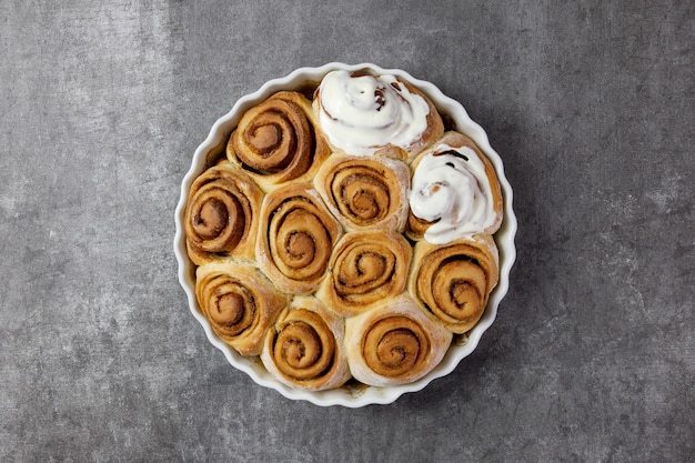 Rollos de canela, bollos de canela en una fuente para hornear con salsa fondant crema de requesón cuajada sobre superficie gris oscuro