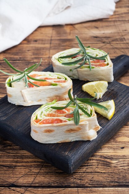 Rollos de pan de pita fino y salmón rojo salado con hojas de lechuga en placa negra sobre mesa de madera