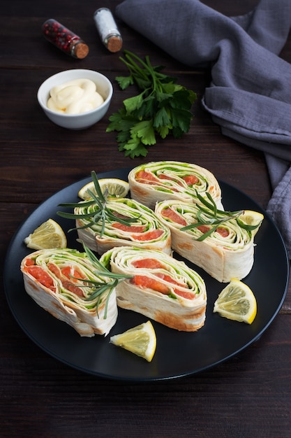 Rollos de pan de pita fino y salmón rojo salado con hojas de lechuga sobre una placa de cerámica negra, mesa de madera oscura.