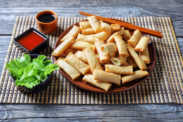 Foto rollos de primavera chinos crujientes del tamaño de un bocado con rellenos de repollo, zanahorias y guisantes verdes en un plato sobre una mesa de madera rústica con salsas en una estera de bambú, vista desde arriba, primer plano