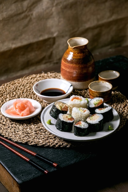 Rollos de sushi caseros con salmón, tortilla japonesa, aguacate, jengibre y salsa de soja con palillos en una servilleta de paja sobre la mesa de madera negra. Juego de sake de bebida de cerámica. Cena al estilo japonés