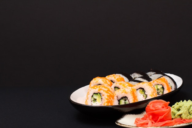 Rollos de sushi con nori, arroz, trozos de aguacate, pepino, huevas de pez volador en plato de cerámica. Plato con jengibre encurtido rojo y wasabi. Fondo negro.