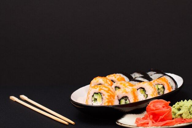 Rollos de sushi con nori, arroz, trozos de aguacate, pepino, huevas de pez volador en plato de cerámica. Plato con jengibre encurtido rojo y wasabi. Fondo negro.