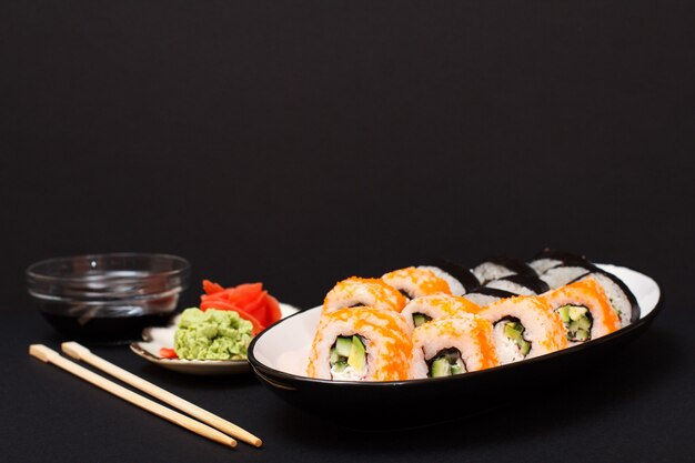 Rollos de sushi con nori, arroz, trozos de aguacate, pepino, huevas de pez volador en plato de cerámica. Plato con jengibre encurtido rojo y wasabi. Recipiente con salsa de soja y palos de madera. Fondo negro.