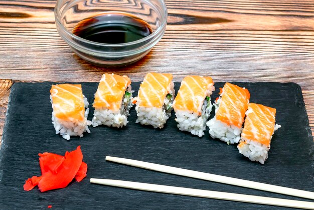 Rollos de sushi de pescado con salsa de soja, jengibre y salmón y palillos en una tabla de cortar negra sobre fondo rústico de madera con espacio para copiar Servicio de comida de mariscos del concepto de restaurante de cerca