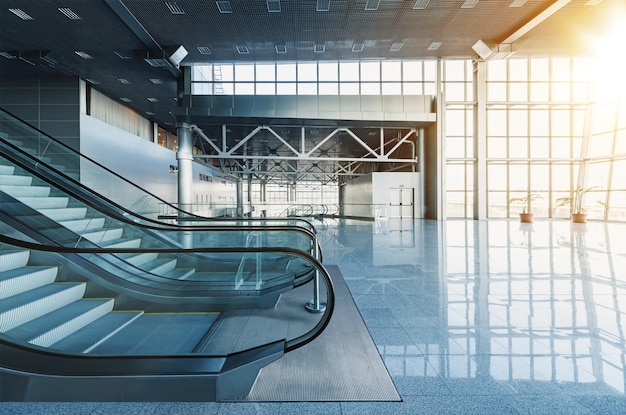 Rolltreppen und Treppen in der Lobby eines modernen Bürogebäudes, Flughafens oder Einkaufszentrums, Glaswände und reflektierender Boden, natürliches Licht und Flare