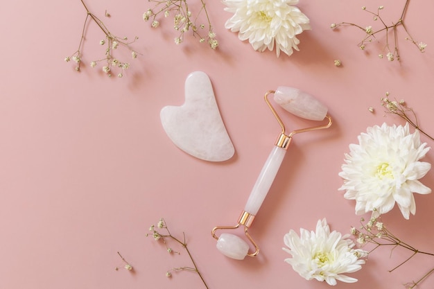 Rolo de massagem facial de quartzo rosa sobre fundo rosa com flores de gipsófila