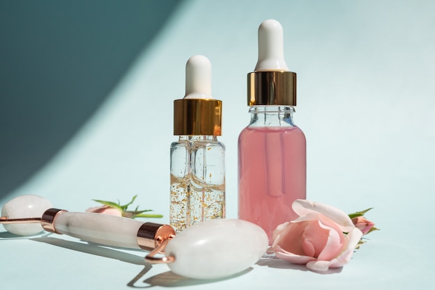 Rolo de massagem facial feito de quartzo rosa com frascos de óleo cosmético ou soro sobre fundo azul. O conceito de cuidados com a pele em casa. Soro ouro 24k e água de rosas para tratamento facial.