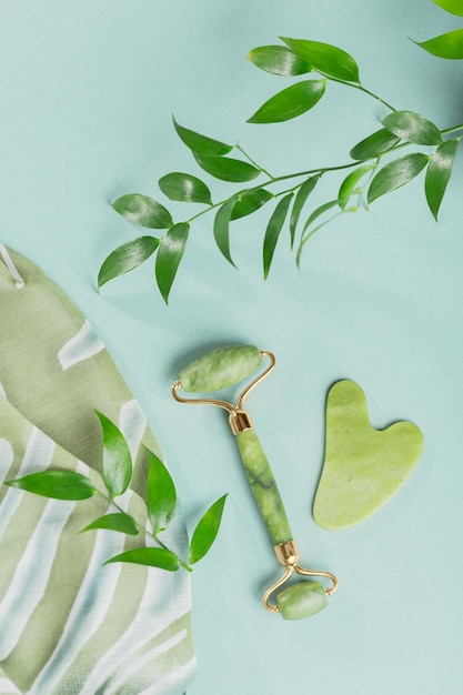 Rolo de massagem para o rosto com duas cabeças e raspador gua sha de pedra jade verde sobre fundo azul claro Massagem facial de beleza SPA conceito de autocuidado