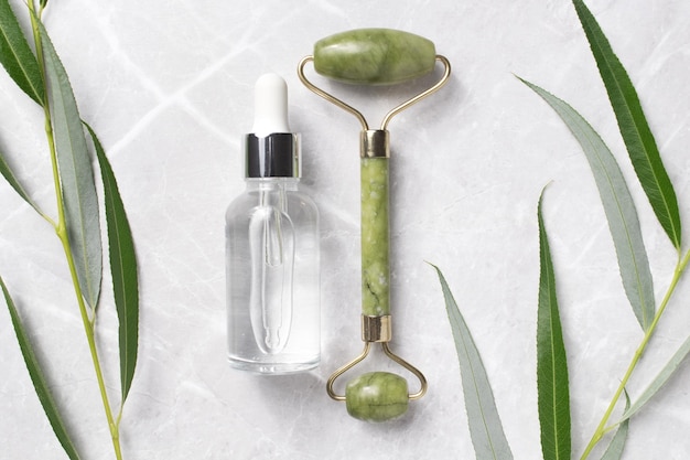 Rolo de rosto de garrafa de soro sem marca com folhas em fundo de mármore Rolo de rosto de jade para terapia de massagem facial Tratamento anti-idade e tonificação