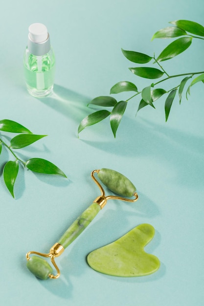 Rolo de rosto de jade verde e raspador de gua sha com folhas em fundo azul claro Ferramenta de massagem para cuidados com a pele facial SPA auto e conceito de tratamento de cuidados de beleza