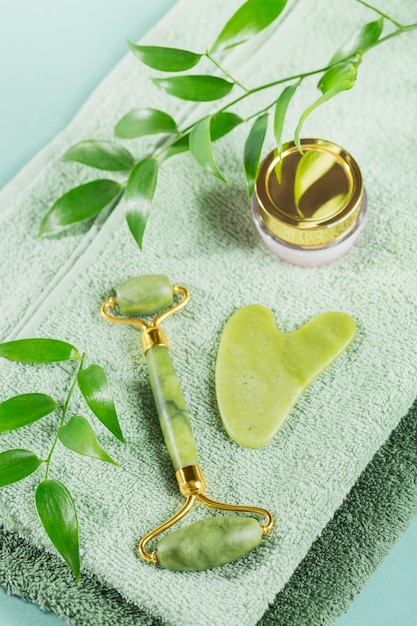 Rolo de rosto de jade verde e raspador de gua sha com folhas em fundo azul claro Ferramenta de massagem para cuidados com a pele facial SPA auto e conceito de tratamento de cuidados de beleza