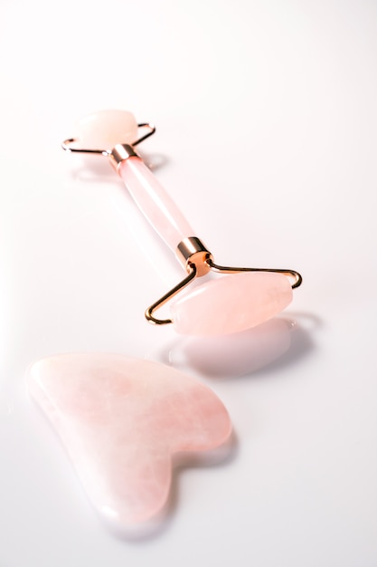 Rolo de rosto rosa e massageador gua sha feito de pedra natural de quartzo sobre fundo branco. Tratamento de levantamento e tonificação em casa.