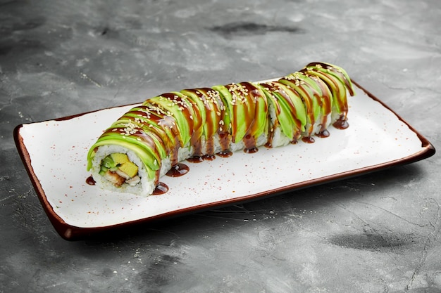 Rolo de sushi clássico dragão verde com molho de abacate, enguia, omelete e unagi em um prato branco sobre uma mesa cinza. Foco seletivo, granulação de ruído na postagem