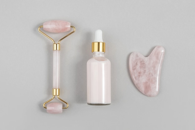 Rolo facial de quartzo rosa cristal, ferramenta de massagem Gua sha e colágeno anti-envelhecimento, soro em frasco de vidro com fundo cinza. Massagem facial para levantamento natural, conceito de beleza Vista superior.