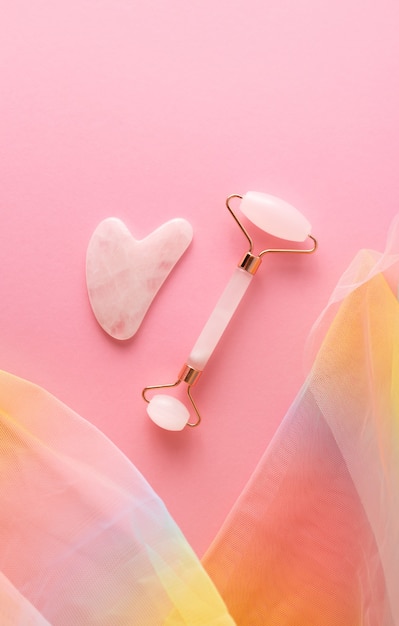 Foto rolo facial de quartzo rosa e gua sha em fundo rosa com tecido de organza colorido arco-íris, vista superior. ferramentas de massageador facial da moda para a pele, vertical