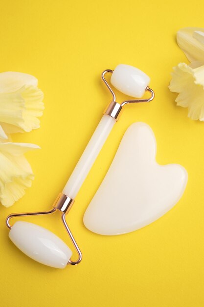 Rolo facial pedra gua sha para massagem facial e flores de narciso em fundo amarelo cuidados cosméticos faciais com a pele e spa conceito de tratamento natural vista superior postura plana