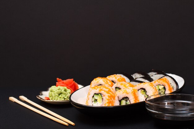 Rolos de sushi com nori, arroz, pedaços de abacate, pepino, ovas de peixe voador no prato de cerâmica. Prato com gengibre em conserva vermelho e wasabi. Tigela com molho de soja. Fundo preto.