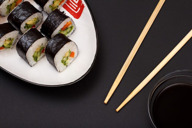 Rolos de sushi em folhas de algas nori com abacate e peixe vermelho no prato de cerâmica. Tigela com molho de soja e palitos de madeira. Vista superior com fundo preto.