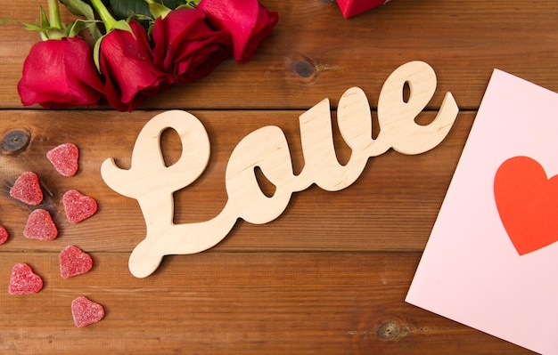 romance, día de san valentín y concepto de vacaciones - cierre de palabra amor, caja de regalo, rosas rojas y tarjeta de saludo con dulces en forma de corazón en madera