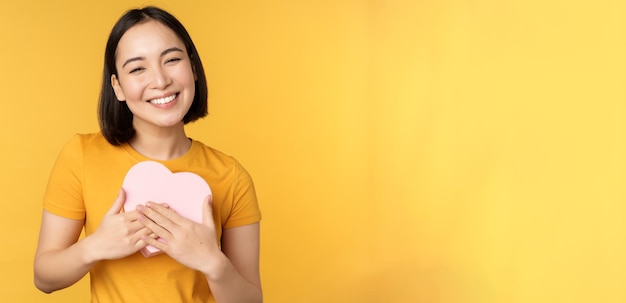 Romance y día de San Valentín Feliz hermosa mujer asiática sosteniendo una gran tarjeta de corazón y sonriendo de pie sobre un fondo amarillo