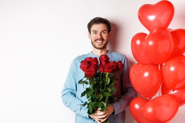 Romance de San Valentín. Hombre joven emocionado con ramo de rosas rojas y globos de corazón sonriendo a la cámara, trae regalos para el amante en la fecha de San Valentín, de pie sobre fondo blanco.