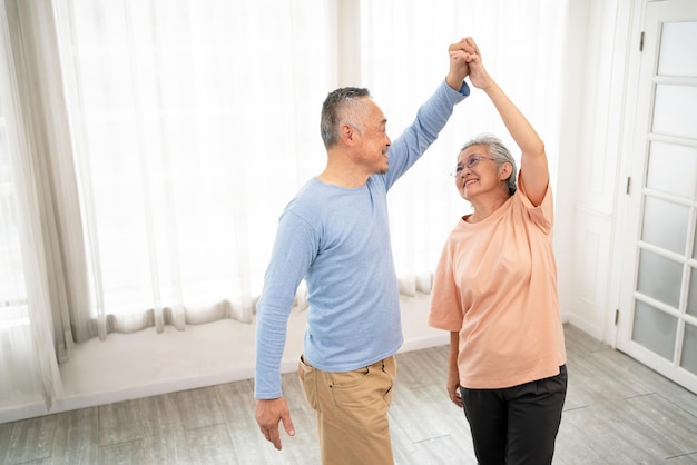 Romántica y alegre pareja de ancianos de bienestar asiático disfruta bailando y tomándose de la mano con música junto con sonrisas y felicidad en la sala de estar Actividad de jubilación para personas mayores en casa