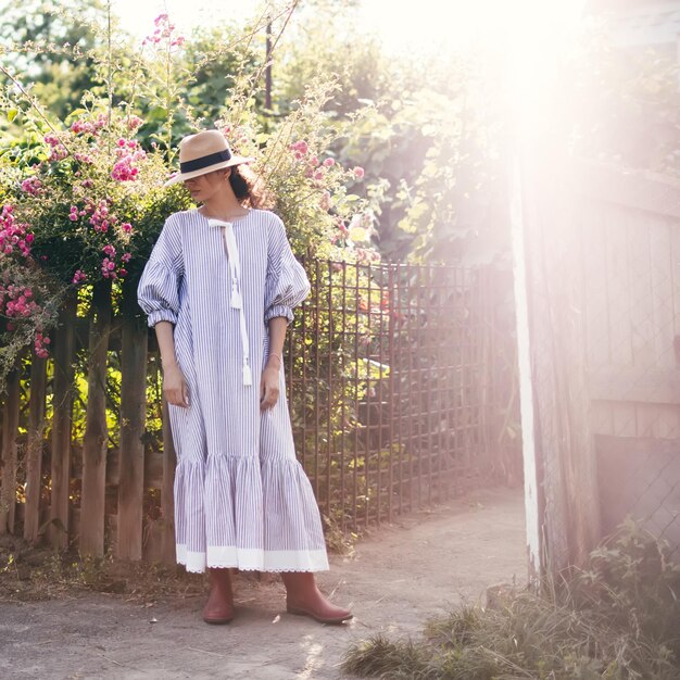 Romántica elegantemente vestida joven adulta hermosa relajándose en un parque rural mirando hacia otro lado Modelo de estilo parisino rubio con un vestido blanco transparente y sombrero de paja caminando contra el jardín