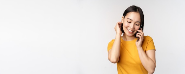 Romántica y linda chica asiática responde llamadas telefónicas hablando por móvil sonriendo coqueta de pie en camiseta amarilla contra fondo blanco