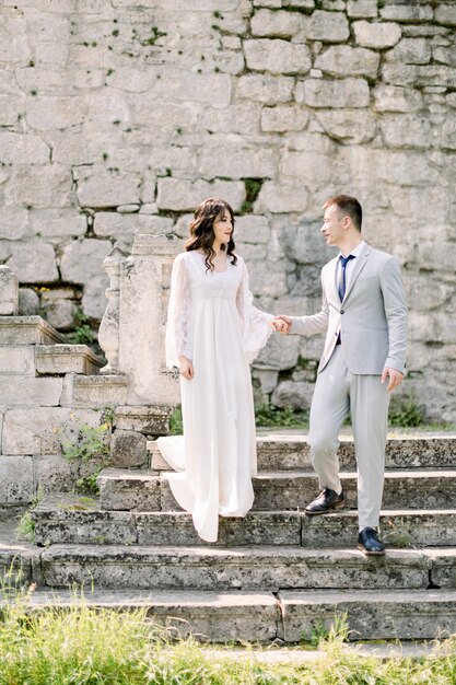 Romántica pareja asiática recién casada posando en las ruinas de un antiguo castillo, cogidos de la mano y de pie en las escaleras de piedra