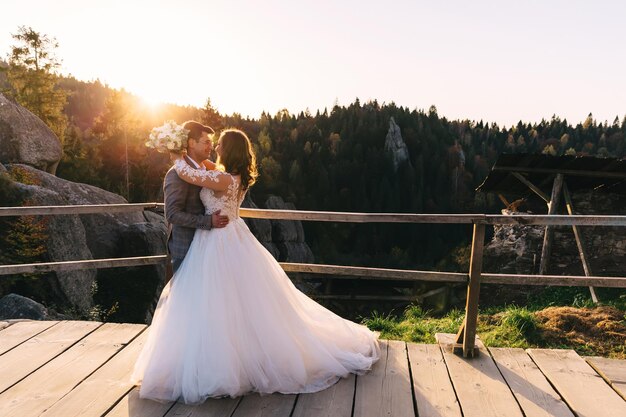 Romántica pareja de cuento de hadas recién casados posando al atardecer sobre un fondo de montañas