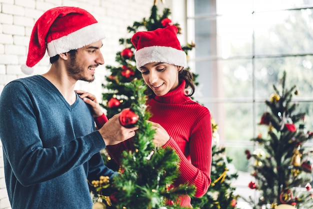 Romántica pareja dulce con sombreros de santa divirtiéndose decorando el árbol de navidad y sonriendo mientras celebra la víspera de año nuevo y disfruta de pasar tiempo juntos en Navidad en casa