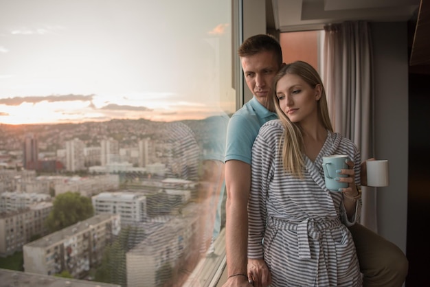 Foto romántica pareja joven feliz disfrutando del café de la noche y hermoso paisaje del atardecer de la ciudad mientras está de pie junto a la ventana