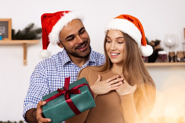 Romántica pareja multiétnica con sombreros de navidad intercambiando regalos